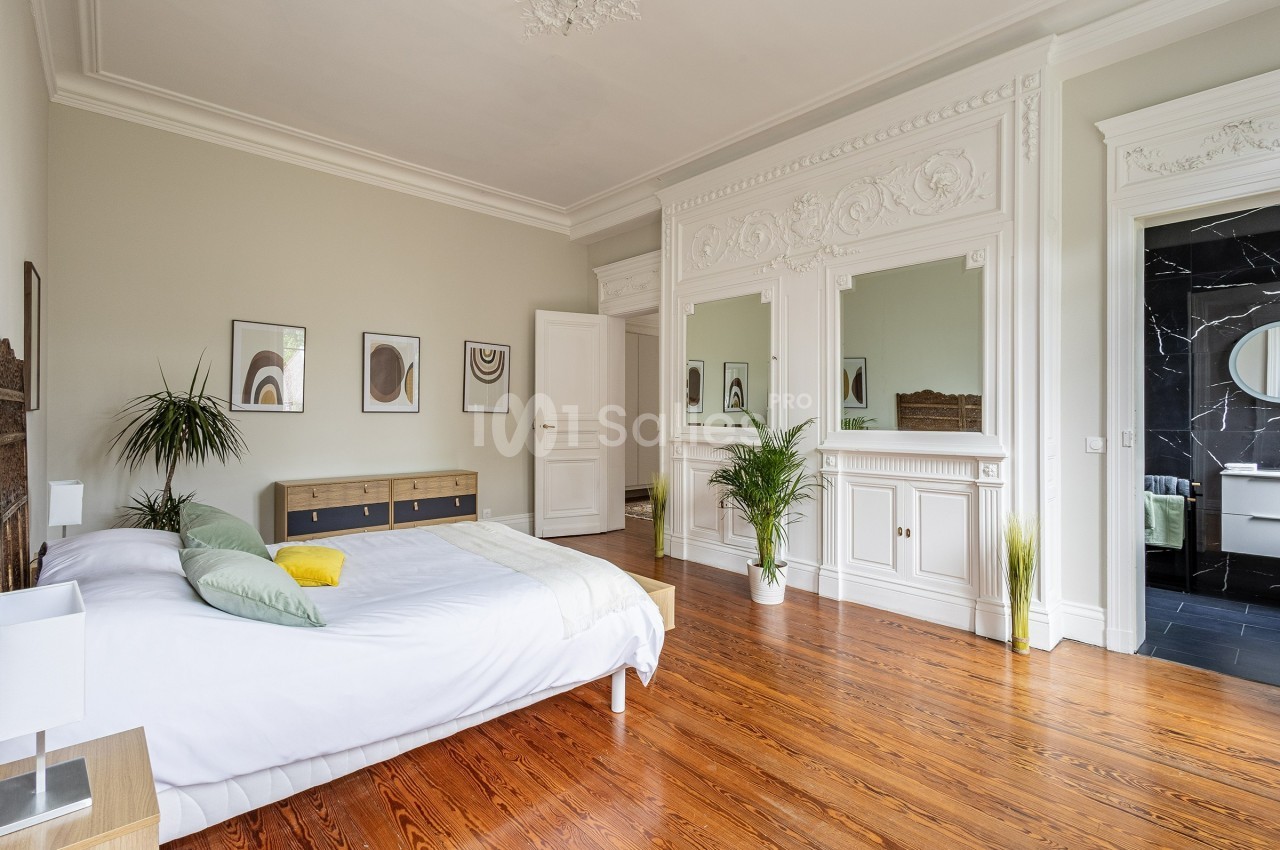 Chambre lumineuse avec parquet en bois, lit double, plantes décoratives et moulures blanches sur les murs.
