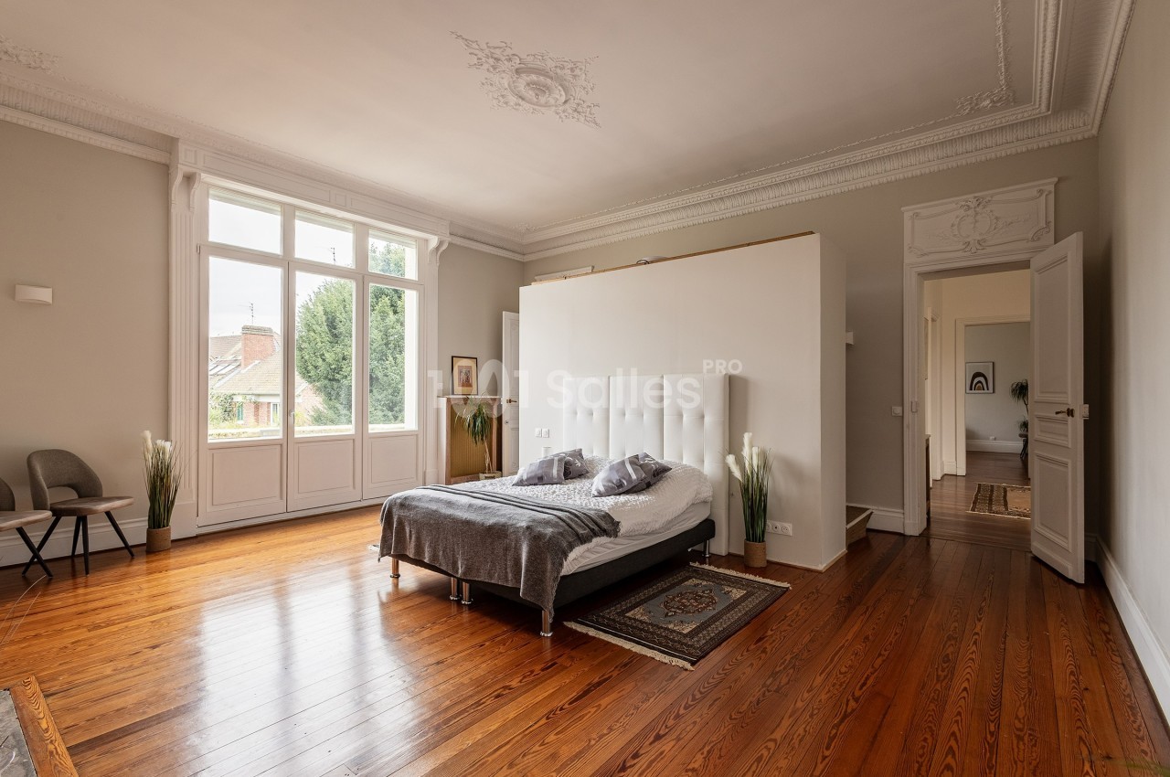 Chambre lumineuse avec parquet en bois, lit double, moulures au plafond et grande fenêtre donnant sur un jardin.