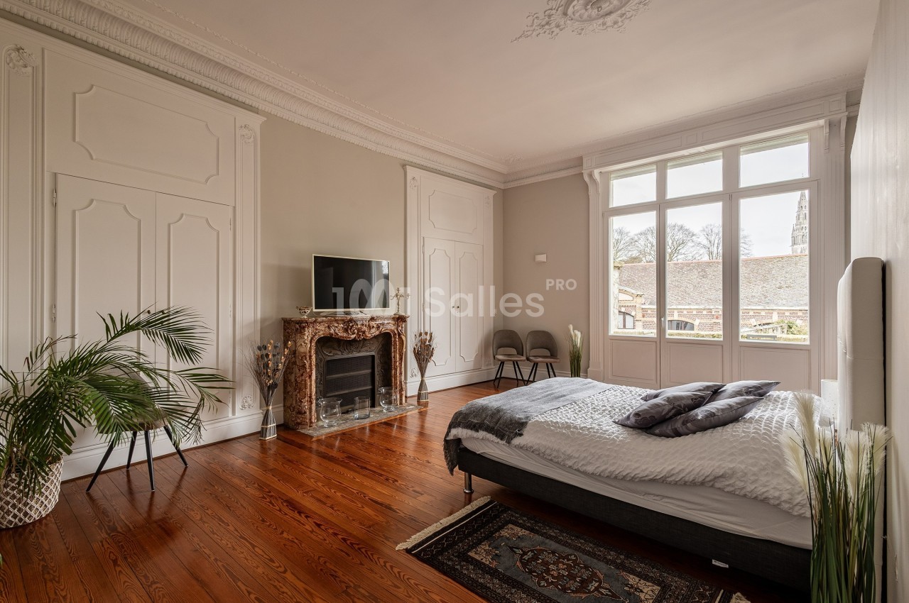 Chambre lumineuse avec parquet, lit double, cheminée en marbre, télévision et grandes fenêtres donnant sur l'extérieur.