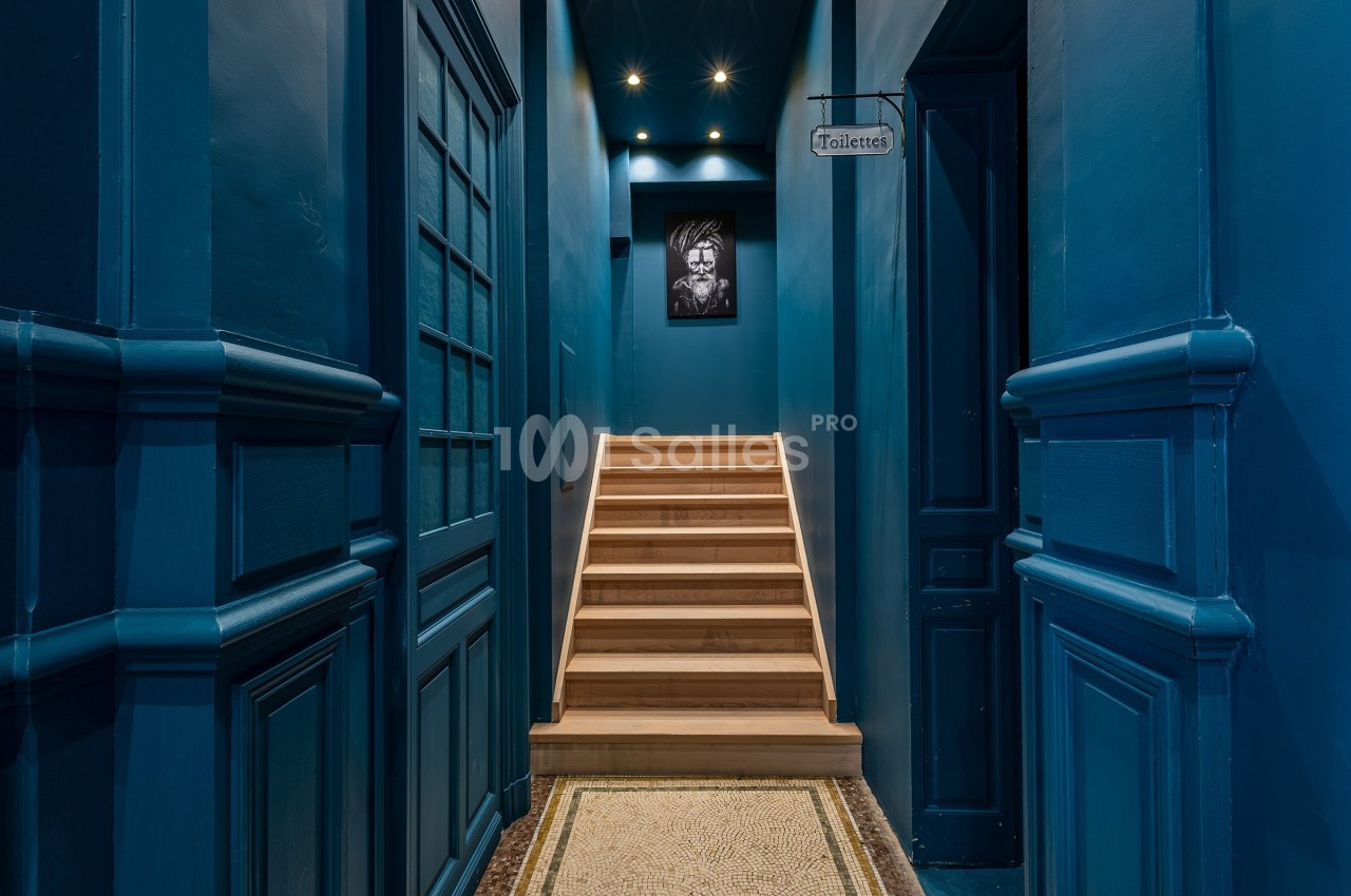 Escalier en bois éclairé menant à un étage, encadré par des murs bleus et un panneau indiquant ’Toilettes’.