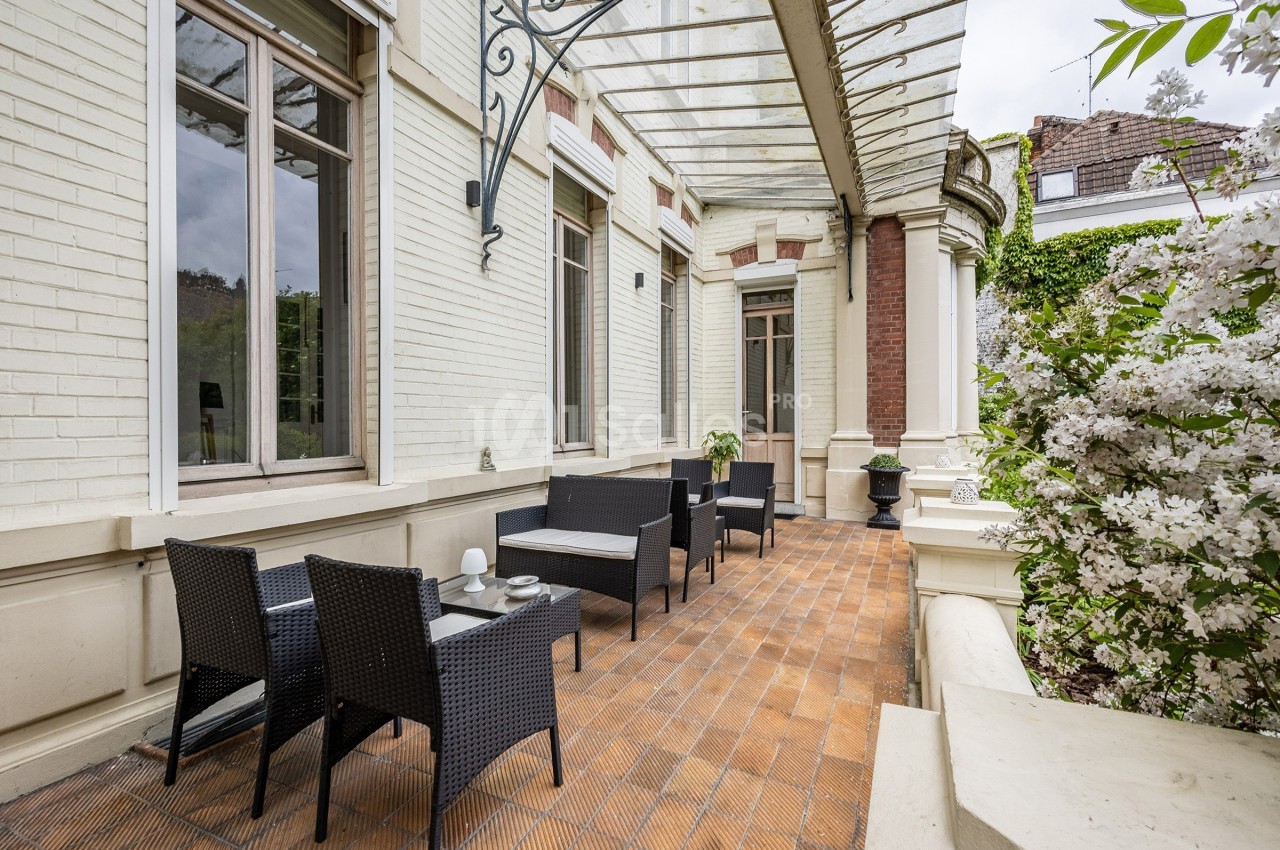 Terrasse en briques avec mobilier en rotin noir, entourée de murs clairs et de végétation fleurie.