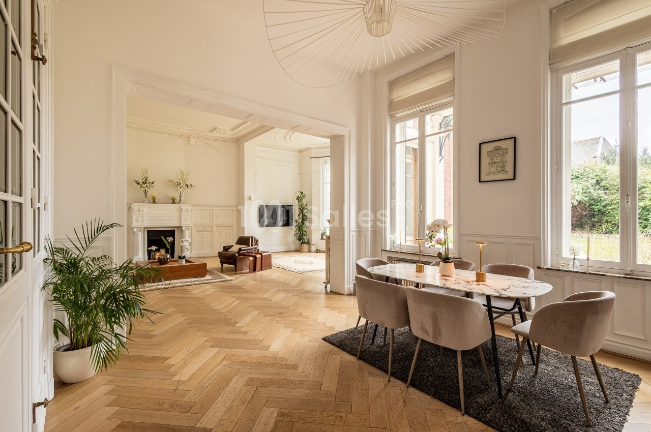 Salle à manger lumineuse avec table, chaises, parquet en bois clair, cheminée et grandes fenêtres donnant sur un jardin.