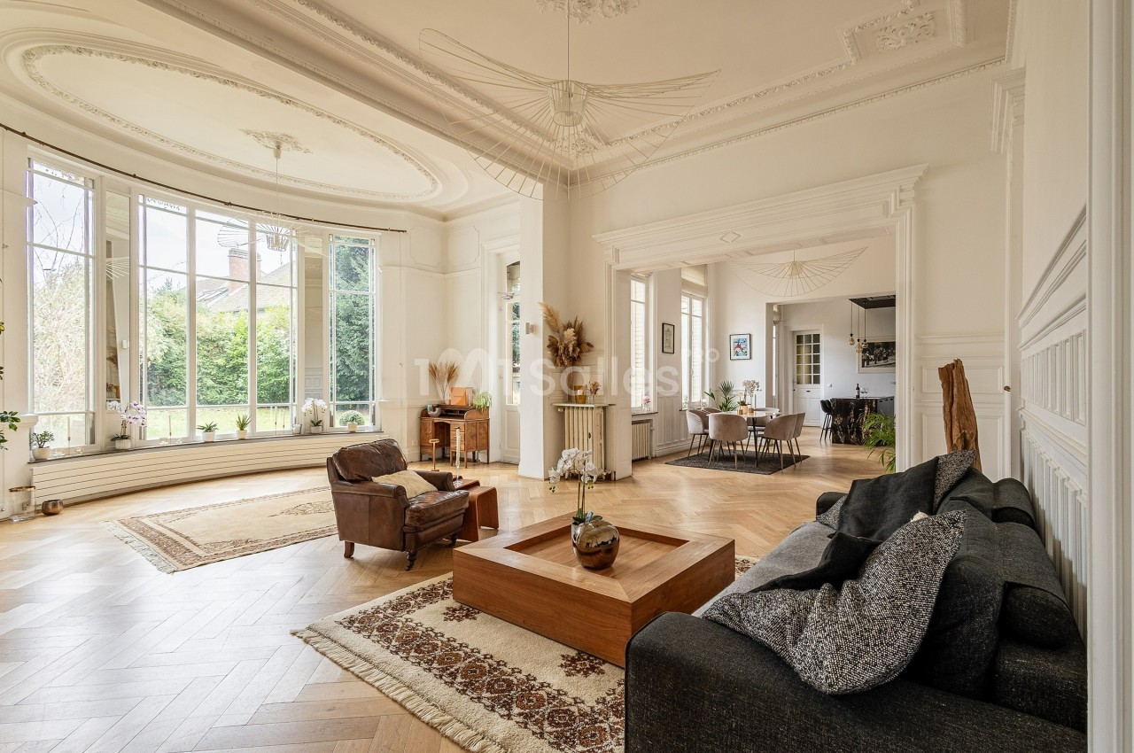 Salon lumineux avec parquet en bois, grandes fenêtres, mobilier élégant et vue sur un jardin verdoyant.