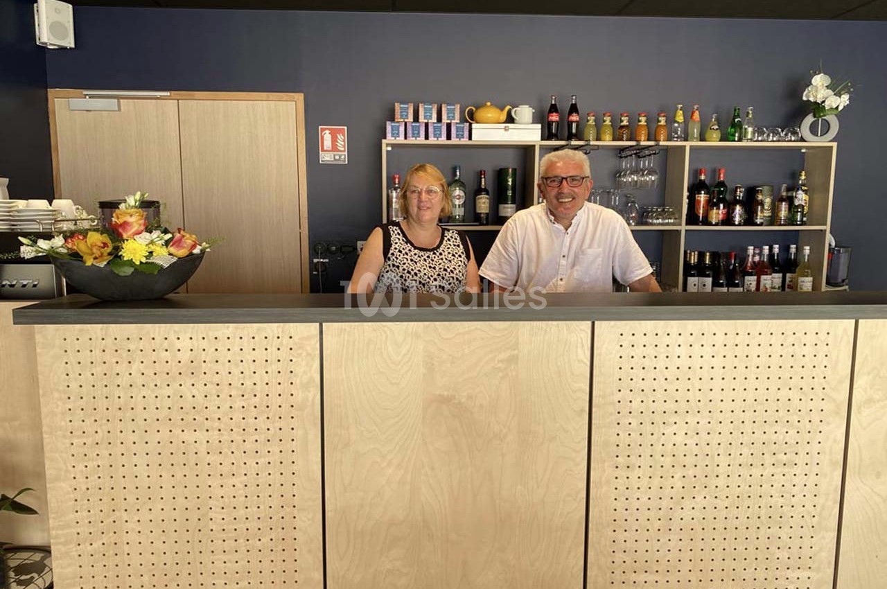 Un homme et une femme souriants derrière un comptoir en bois, avec des étagères de boissons en arrière-plan.