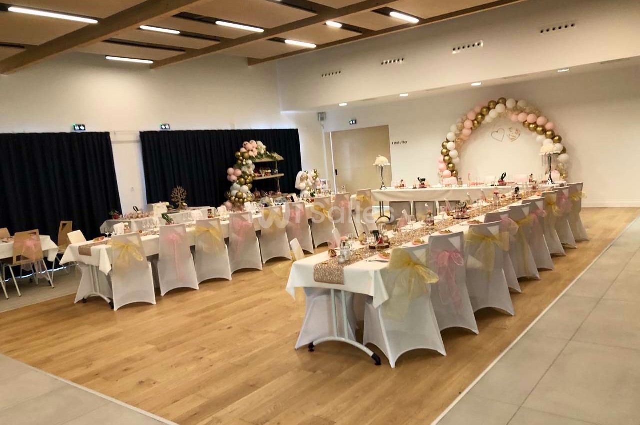 Salle décorée pour une réception avec tables alignées, nappes blanches, chaises ornées de nœuds et arche de ballons.
