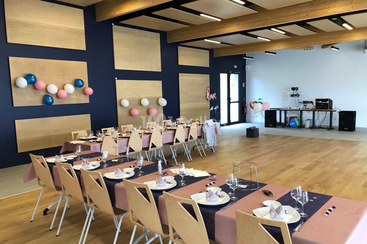 Salle décorée pour un événement, avec tables dressées, nappes roses et bleues, et ballons accrochés aux murs.