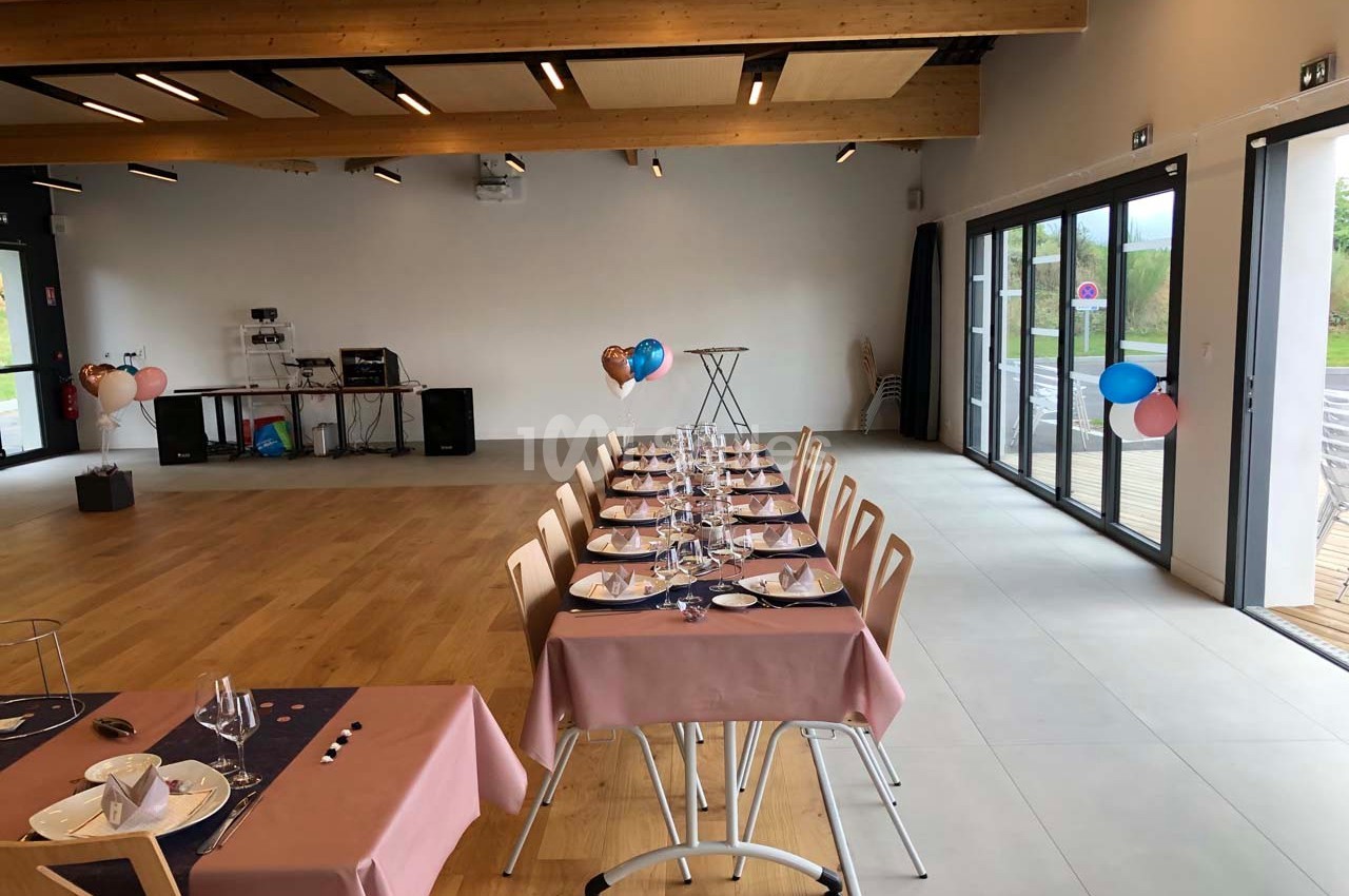 Salle décorée pour un événement, avec tables dressées, nappes roses et ballons, éclairée par des baies vitrées.