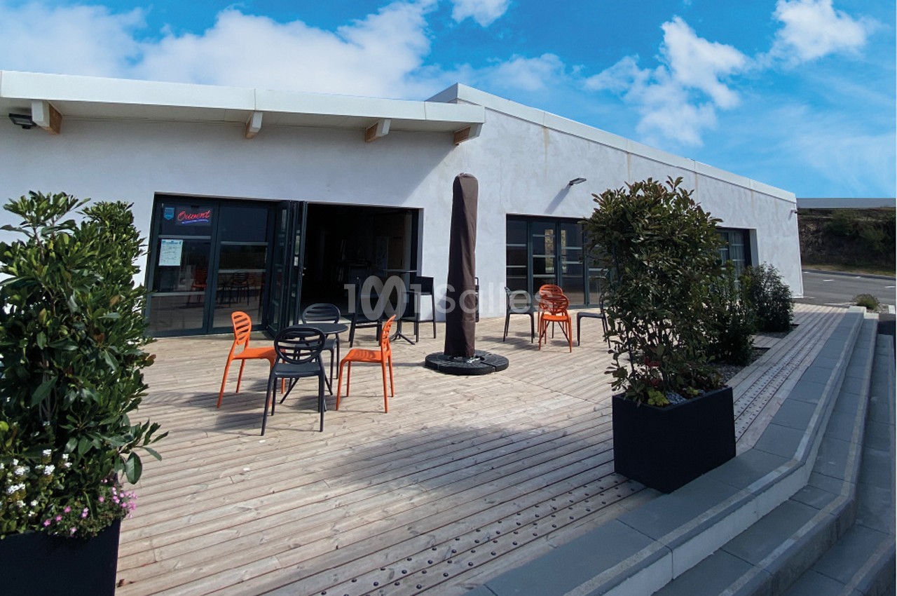 Terrasse en bois avec tables et chaises colorées, entourée de plantes, devant un bâtiment moderne sous un ciel bleu.