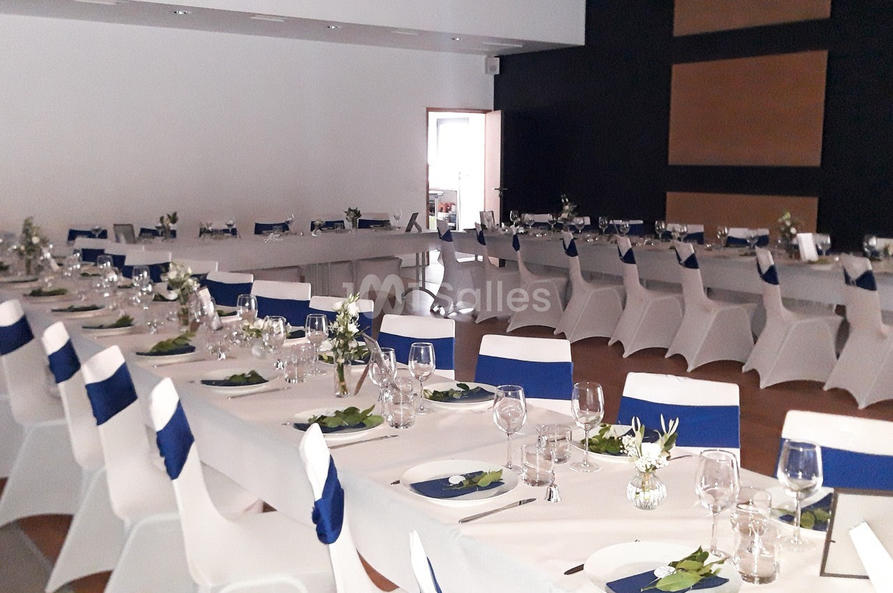 Salle de réception décorée avec des tables dressées, nappes blanches, serviettes bleues et arrangements floraux.