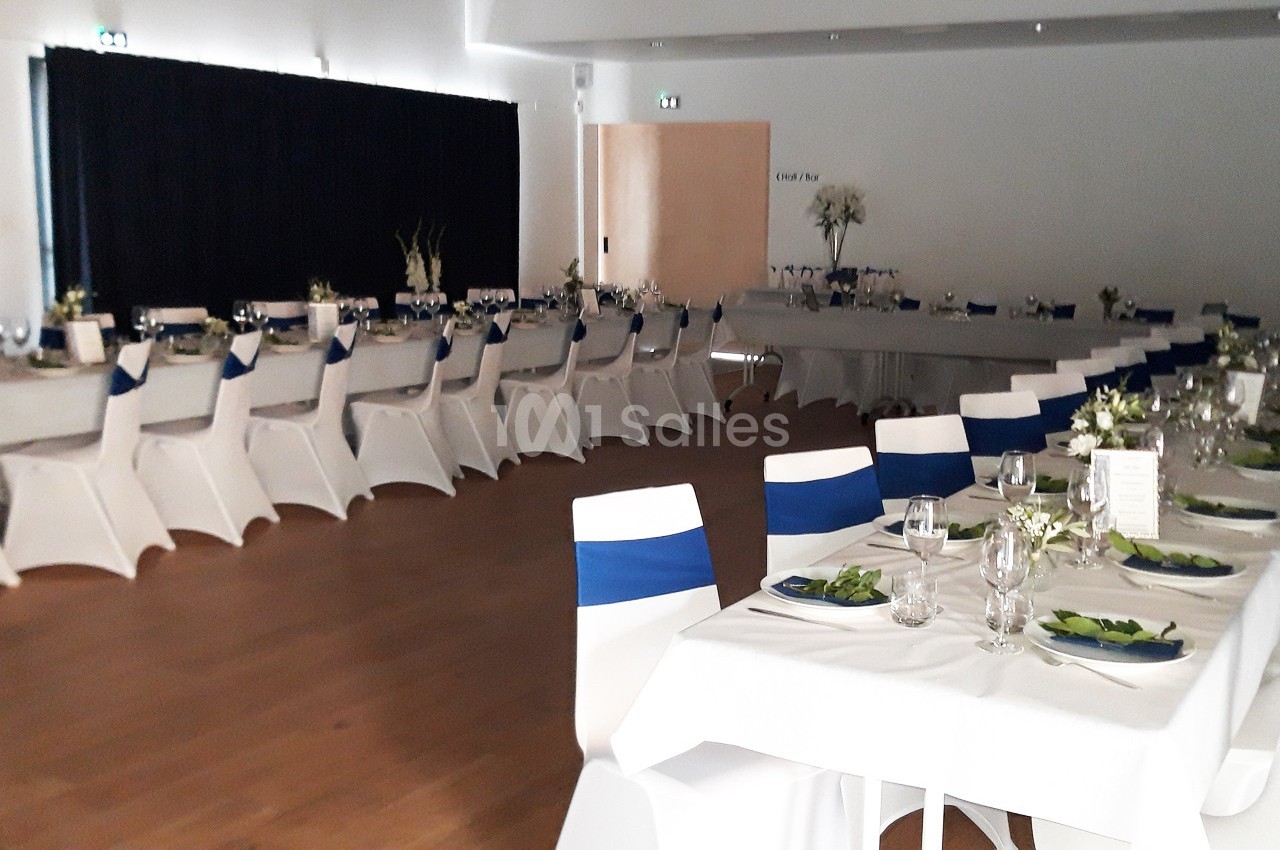 Salle de réception avec tables dressées, nappes blanches, chaises décorées de rubans bleus et arrangements floraux.