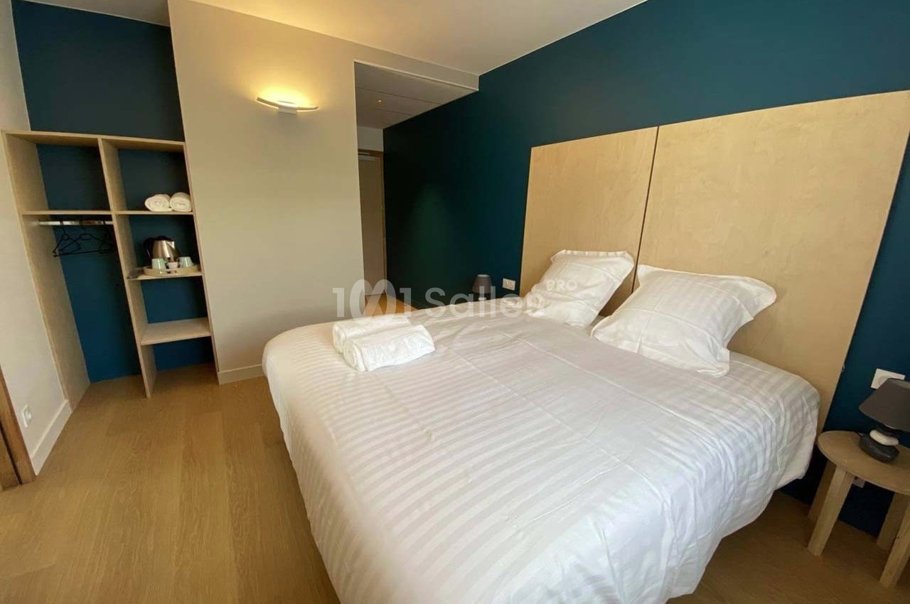 Lit double avec draps blancs dans une chambre moderne, mur bleu, éclairage doux et espace de rangement ouvert.