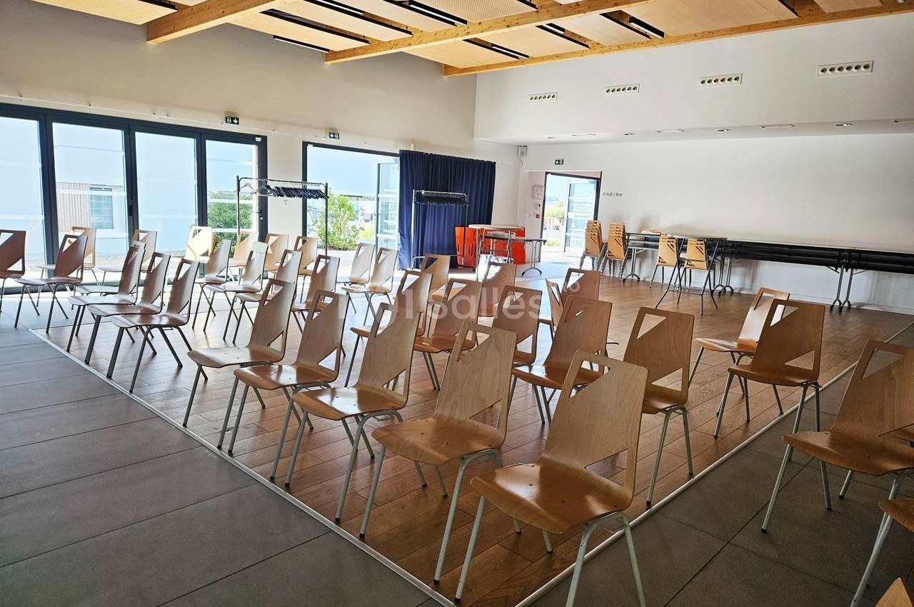 Salle lumineuse avec des rangées de chaises en bois vides, grandes baies vitrées et plafond avec poutres apparentes.