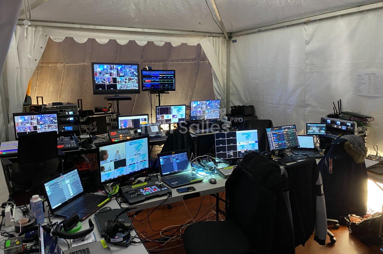 Poste de contrôle audiovisuel sous une tente, avec plusieurs écrans, ordinateurs et équipements techniques.