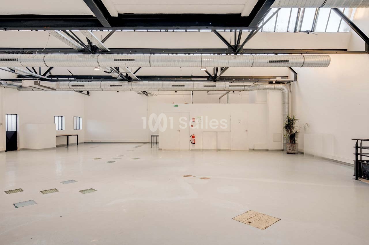 Intérieur d'un grand espace industriel lumineux avec plafond en poutres métalliques, conduits apparents et murs blancs.