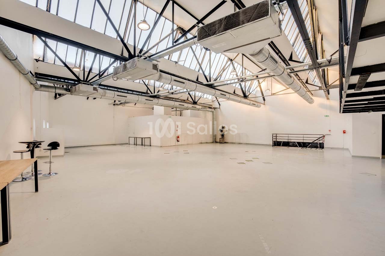Intérieur d'un grand espace industriel lumineux avec plafond en verrière, poutres métalliques et sol en béton clair.