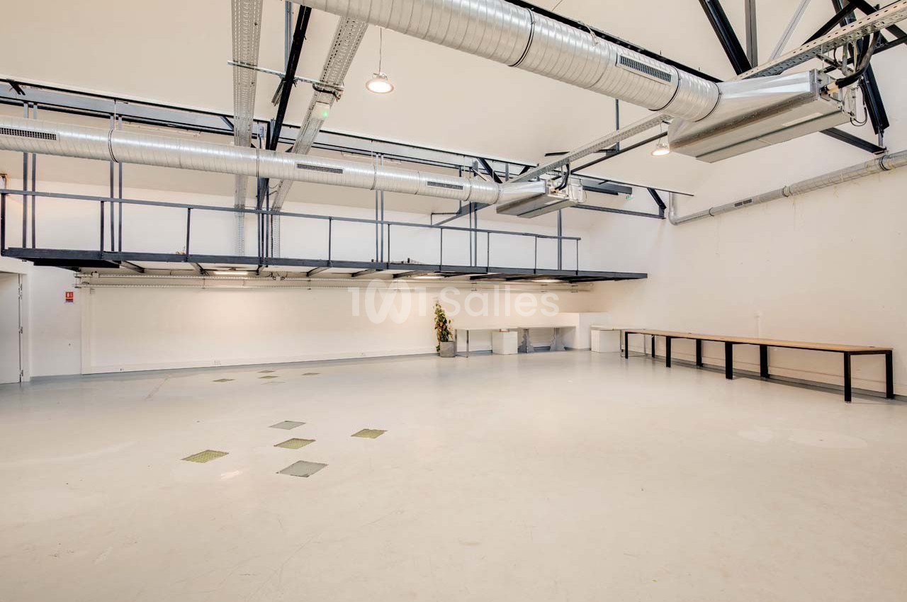 Grand espace intérieur lumineux avec sol clair, plafond haut, conduits apparents et mezzanine métallique.