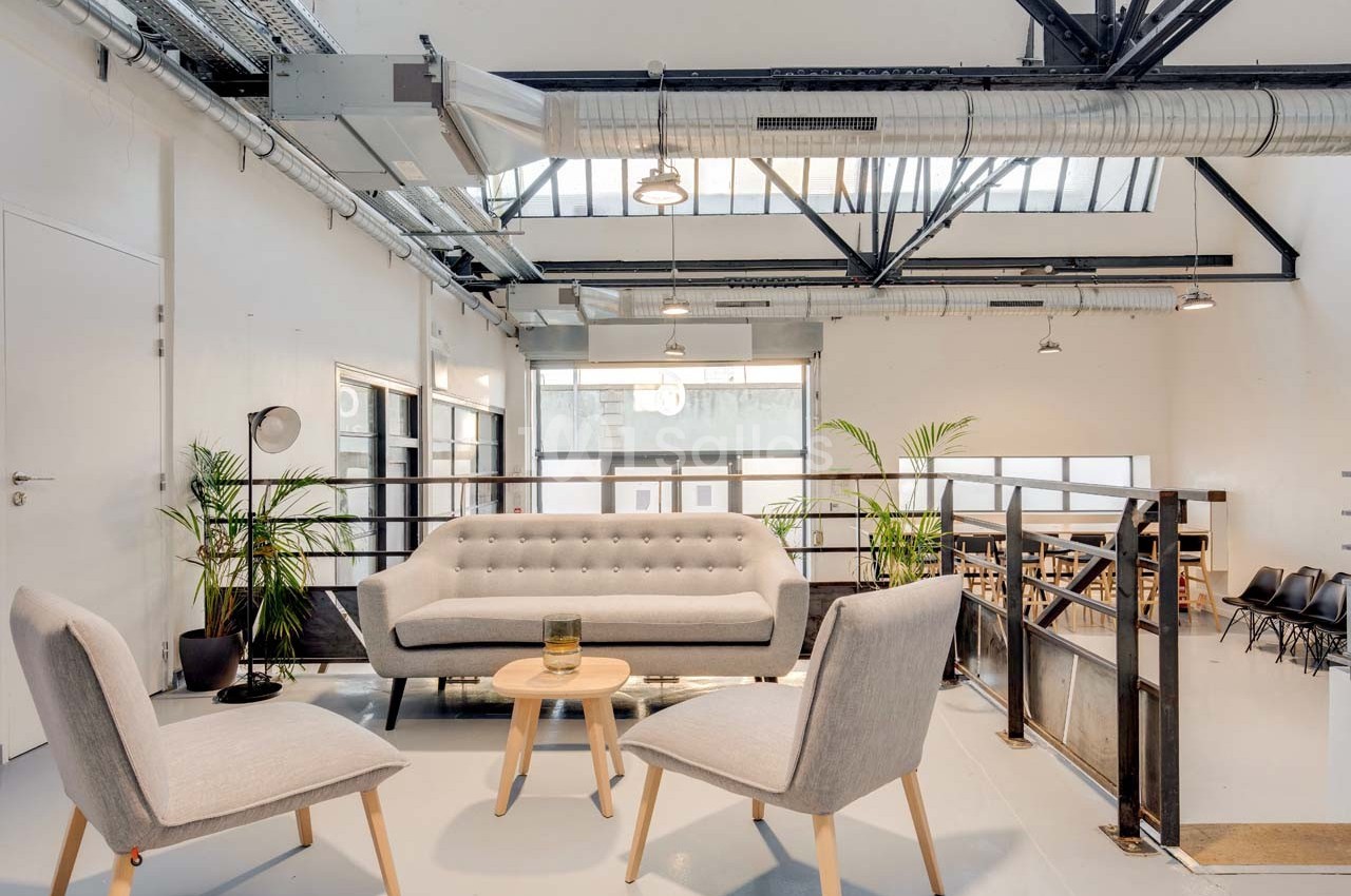 Espace lumineux avec canapé, fauteuils, plantes et mezzanine, dans un intérieur industriel avec verrière et poutres…