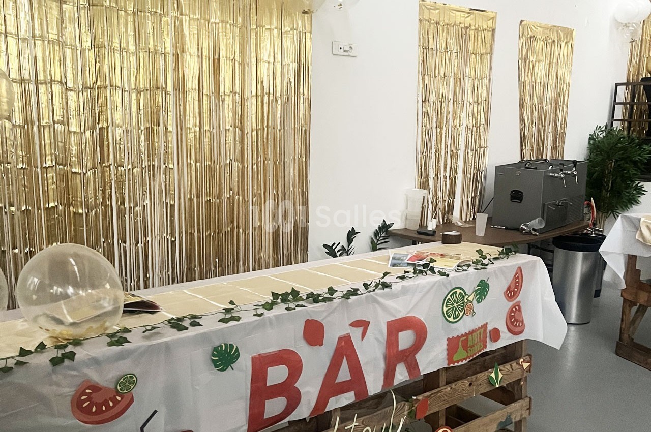 Table de bar décorée avec une nappe blanche, des motifs de fruits et des guirlandes, devant un fond doré scintillant.