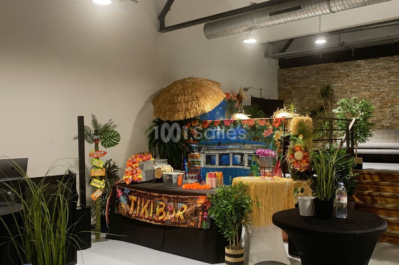 Stand de bar tropical décoré avec des plantes, des fleurs colorées, une enseigne ’Tiki Bar’ et des accessoires exotiques.