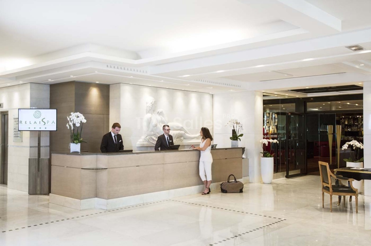 Réception d'un hôtel avec deux employés accueillant une cliente dans un hall lumineux et moderne.