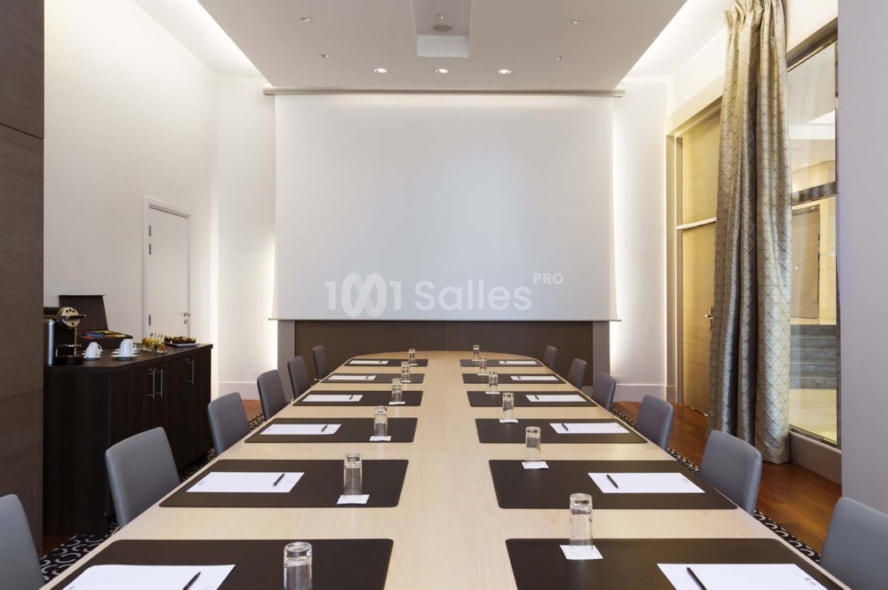 Salle de réunion moderne avec une grande table rectangulaire, chaises grises et écran de projection au fond.