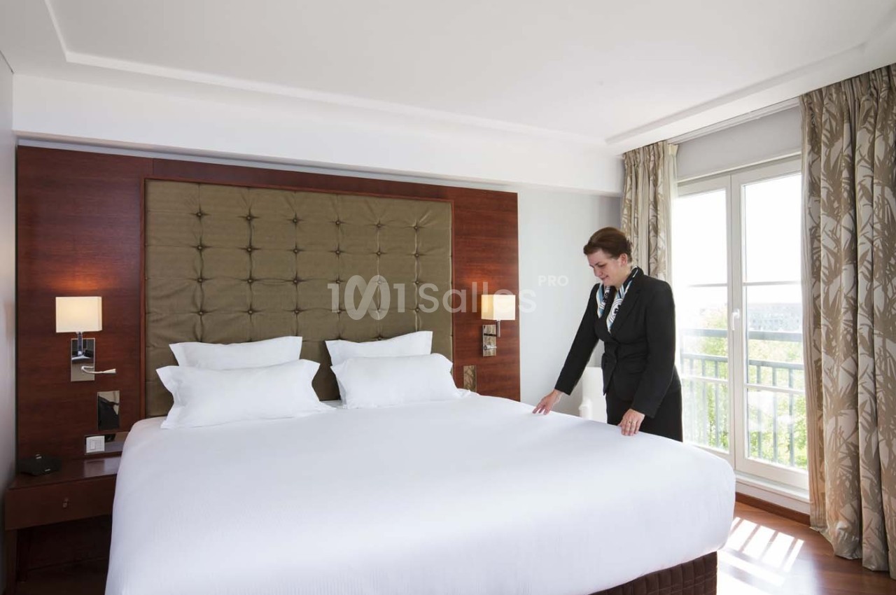 Une femme ajuste les draps d'un grand lit dans une chambre lumineuse avec des rideaux et une grande fenêtre.