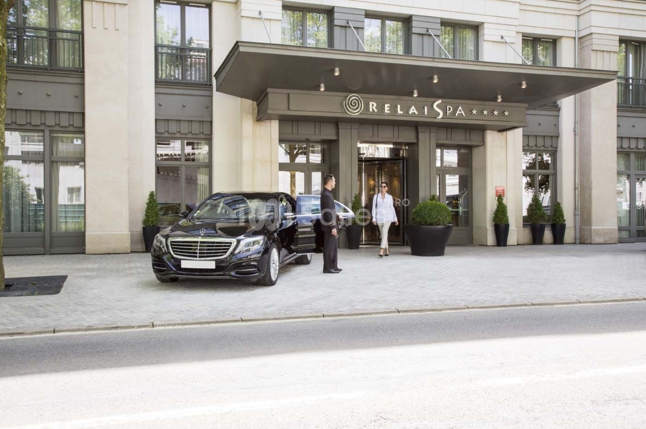Entrée d'un hôtel avec une voiture noire garée devant, un chauffeur ouvrant la porte pour une cliente.