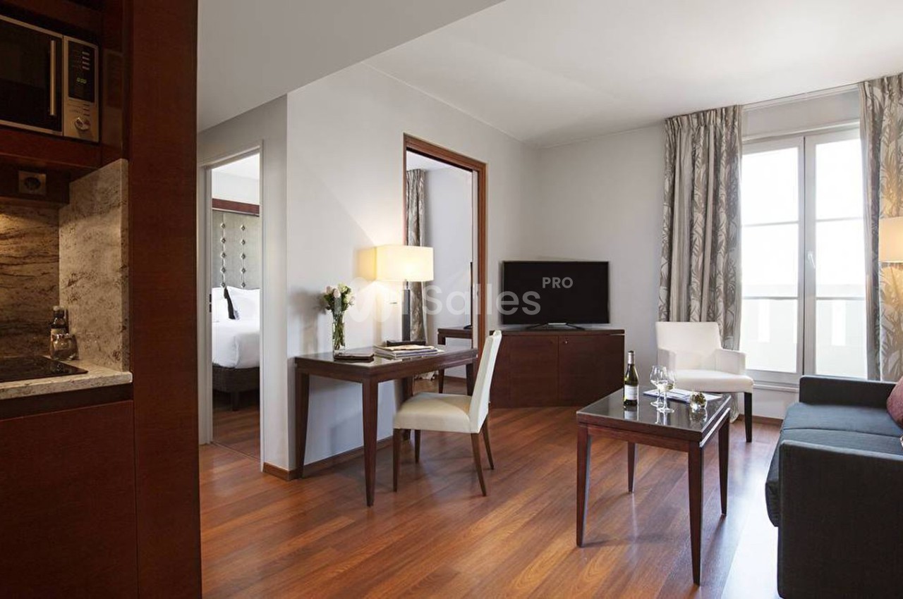 Salon d'un appartement avec parquet, canapé, table, télévision, cuisine ouverte et chambre visible en arrière-plan.