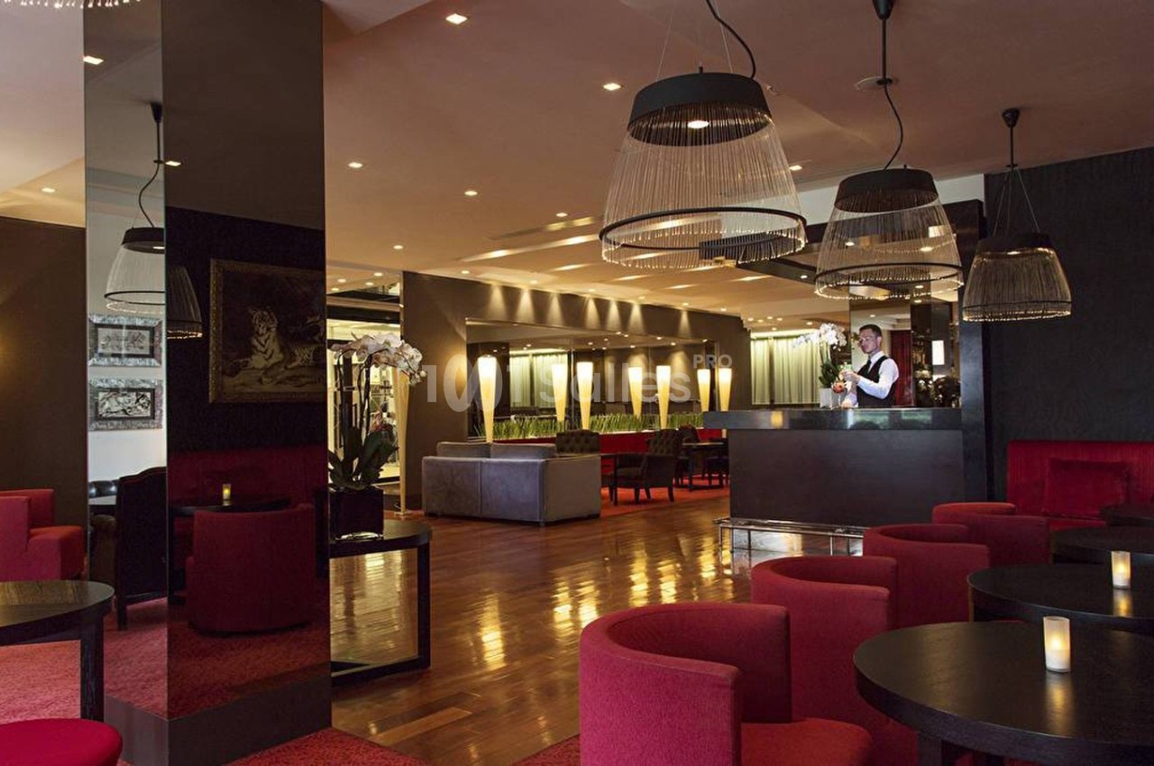 Salle de restaurant élégante avec tables noires, fauteuils rouges, éclairage tamisé et un serveur au comptoir.