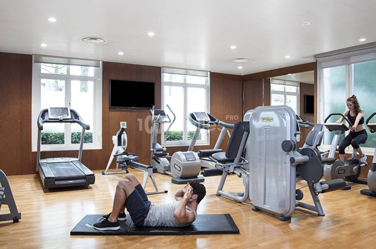 Salle de sport lumineuse avec équipements de cardio et musculation, un homme fait des abdominaux au sol.
