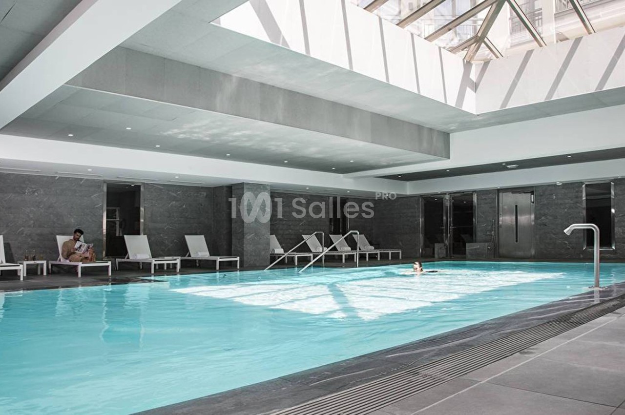 Piscine intérieure lumineuse entourée de chaises longues, avec un plafond en verre laissant entrer la lumière naturelle.
