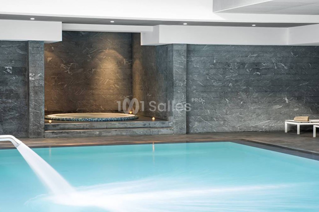 Piscine intérieure avec cascade d'eau, jacuzzi encastré dans un mur en pierre et transat blanc sur le côté.
