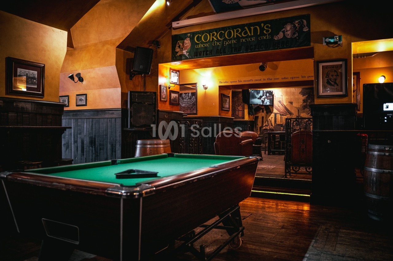 Table de billard dans un bar à l'éclairage tamisé, entourée de décorations murales et de tonneaux en bois.