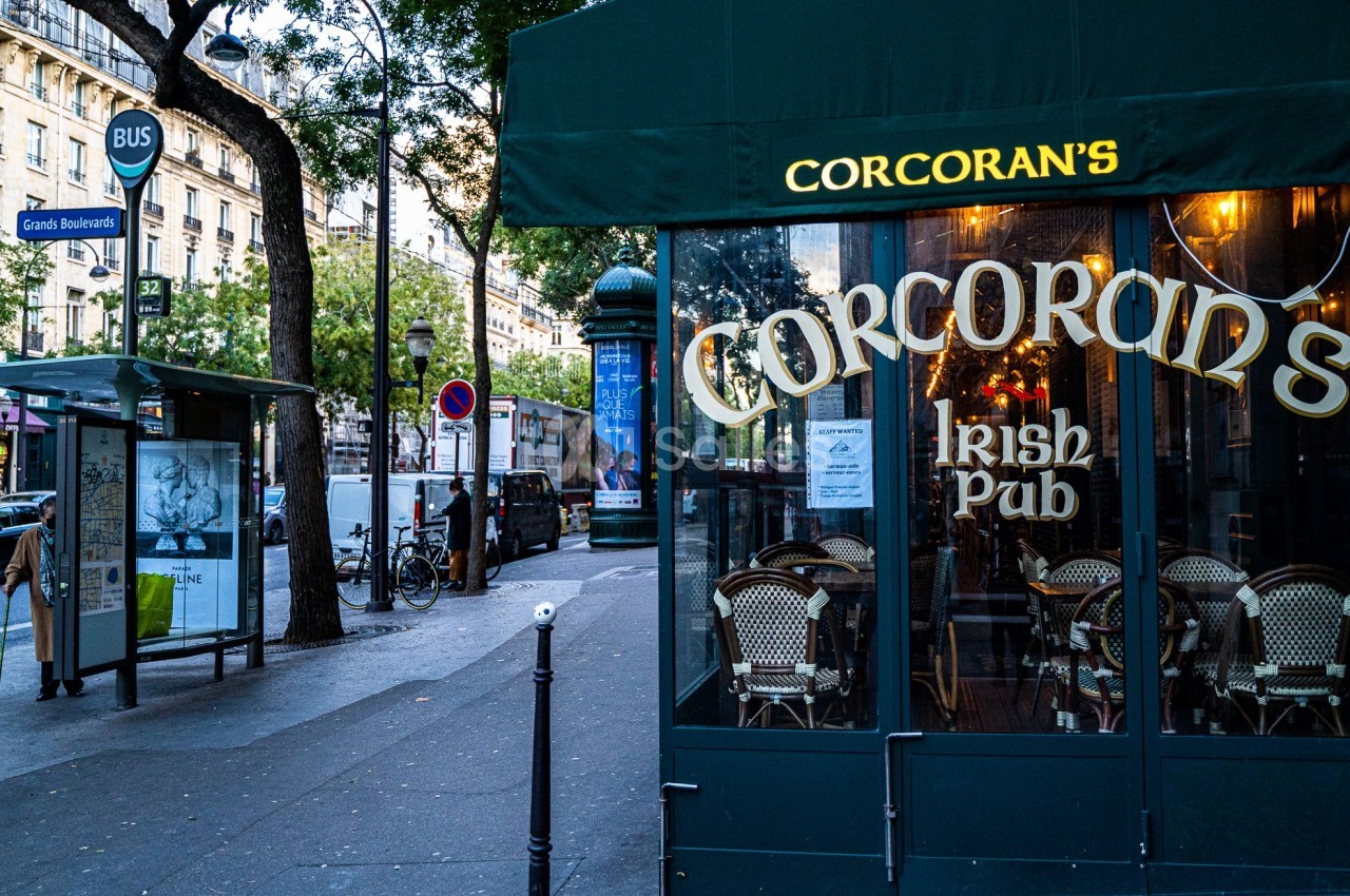 Façade d'un pub irlandais avec terrasse, situé près d'un arrêt de bus dans une rue urbaine animée.