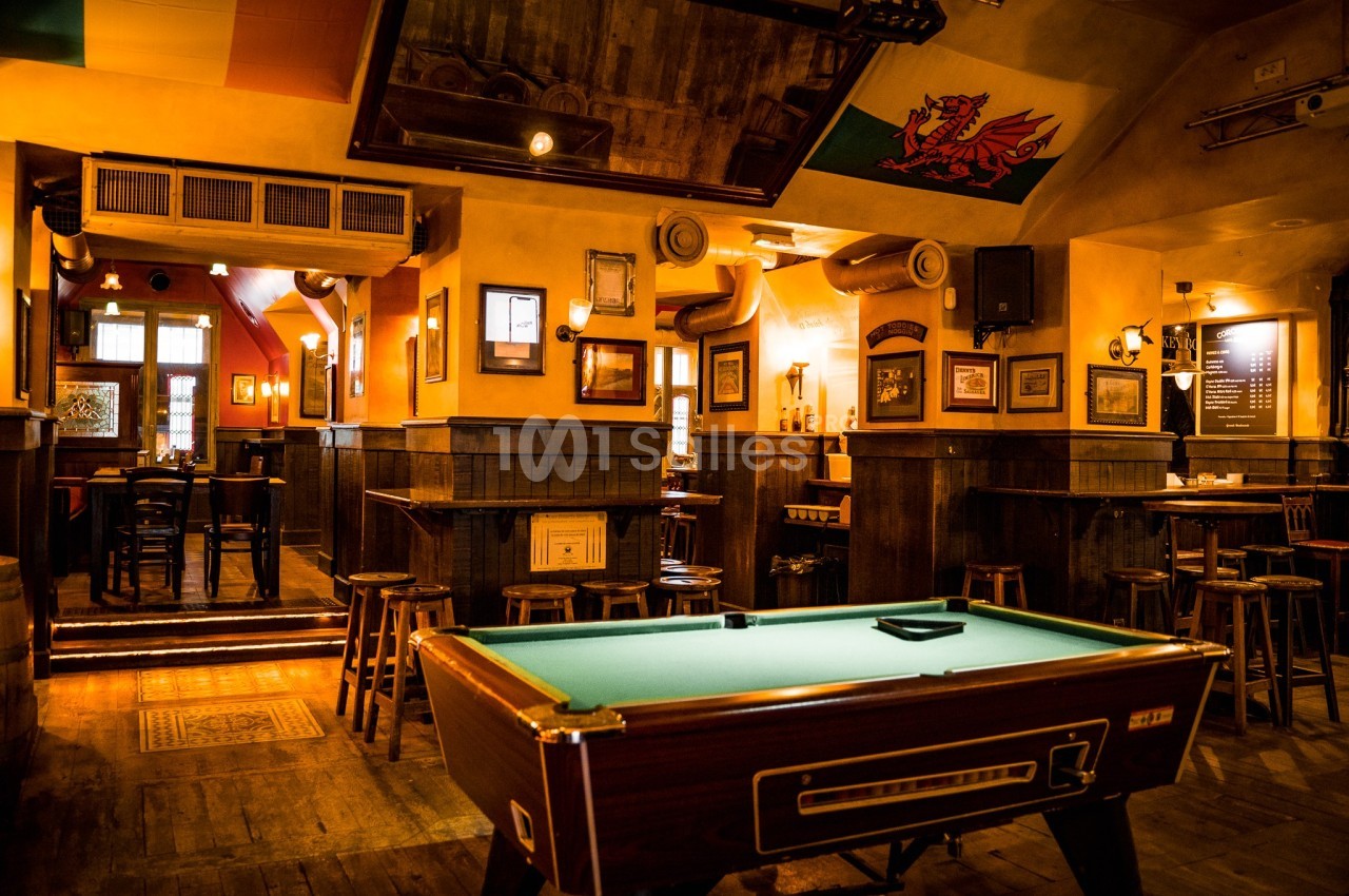 Salle de bar chaleureuse avec tables hautes, tabourets en bois et une table de billard au premier plan.