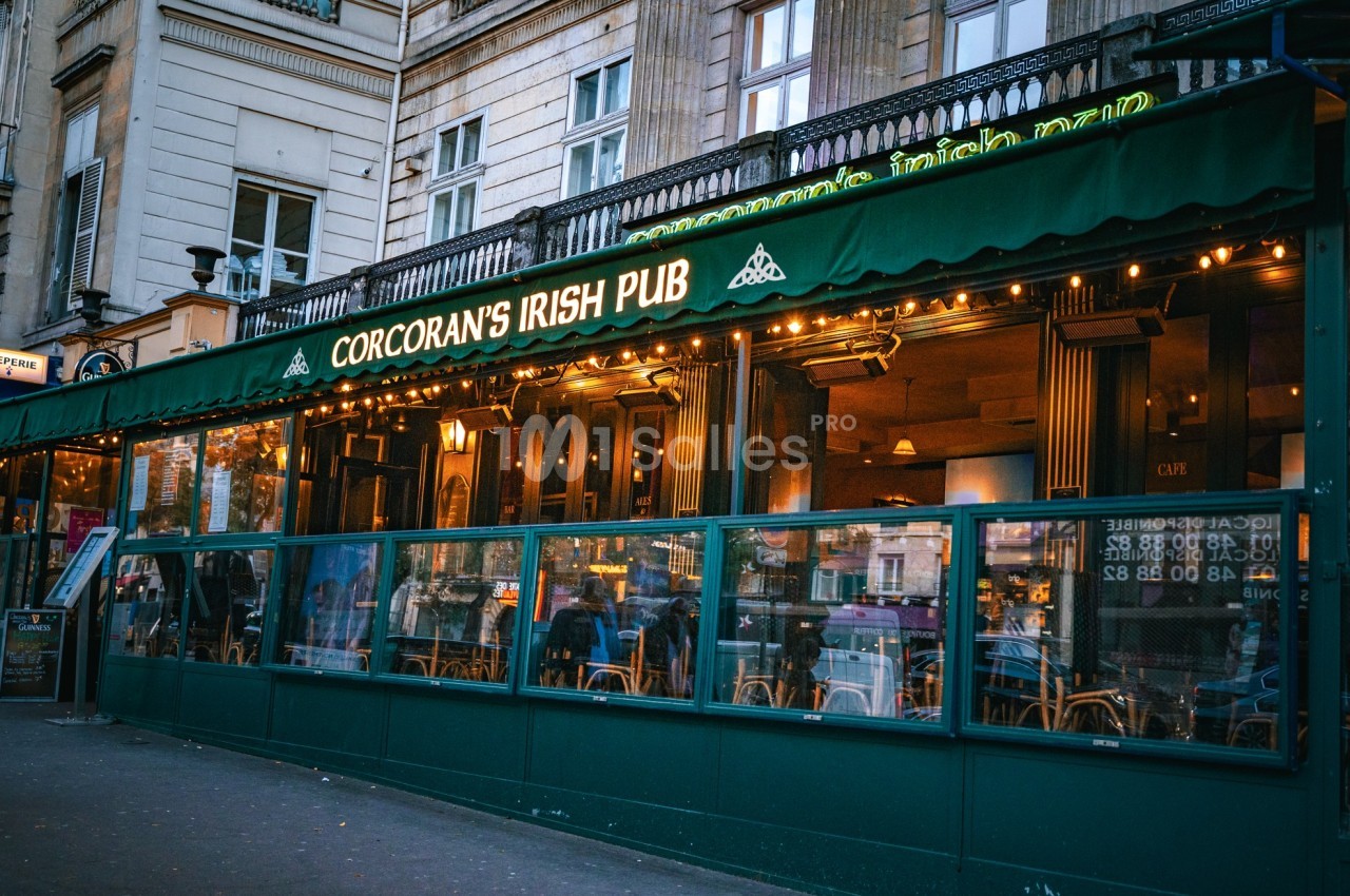 Façade d'un pub irlandais avec terrasse vitrée, enseigne lumineuse et décorations vertes, situé en milieu urbain.