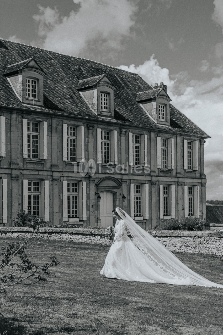 Une mariée en robe blanche marche sur une pelouse devant un grand bâtiment ancien en pierre.