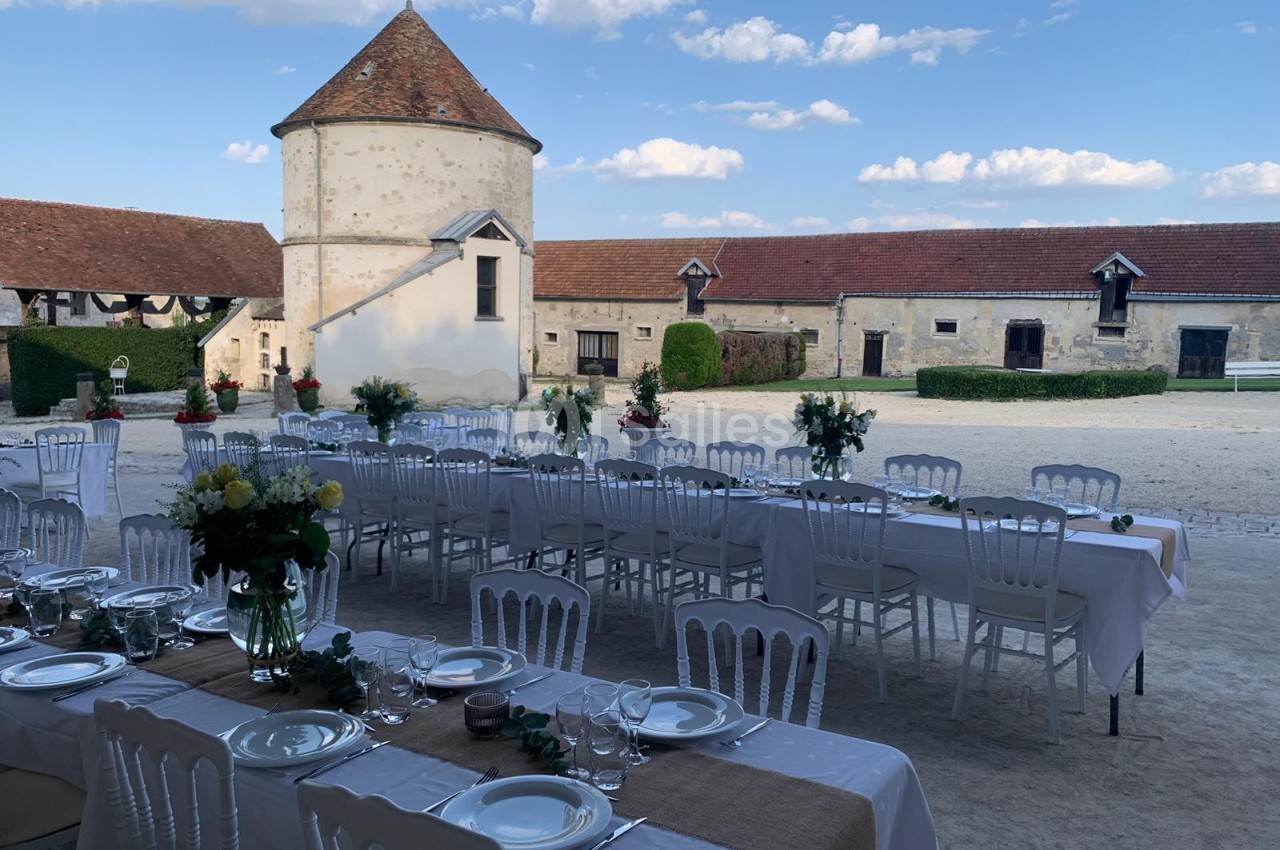 Tables dressées avec nappes blanches et décorations florales dans une cour de ferme avec bâtiments en pierre et ciel dégagé.