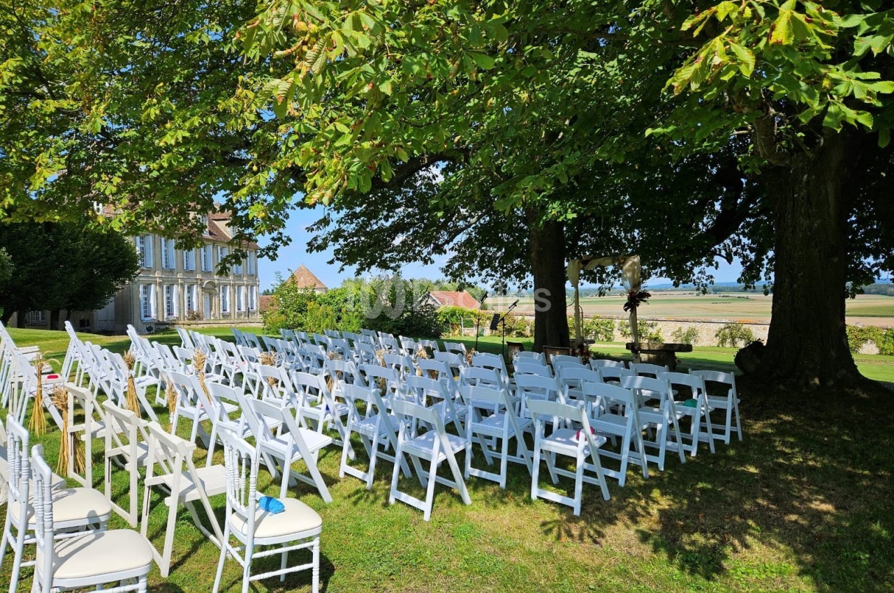 Chaises blanches alignées en extérieur sous des arbres, préparées pour une cérémonie avec vue sur un paysage champêtre.