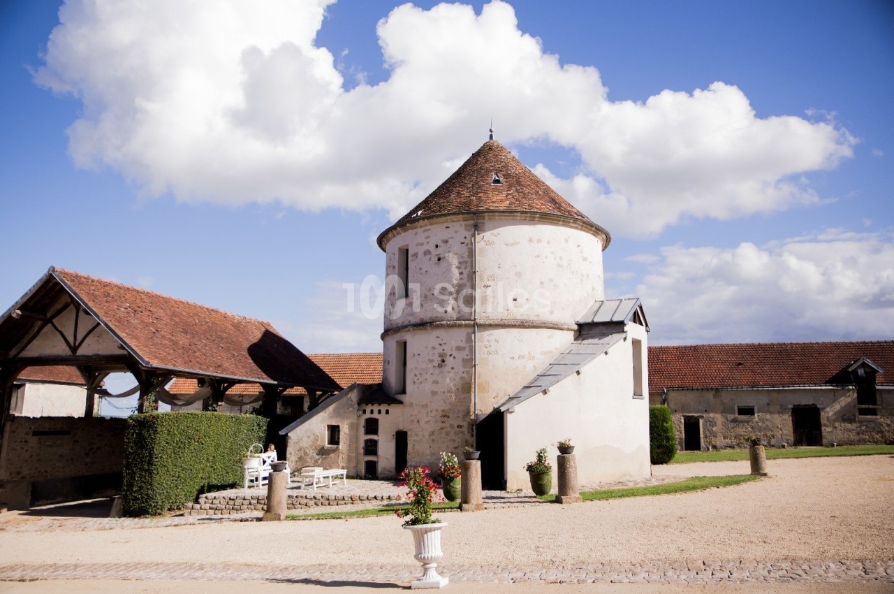 Bâtiment en pierre avec toit en tuiles, entouré de dépendances et d'une cour gravillonnée sous un ciel bleu.
