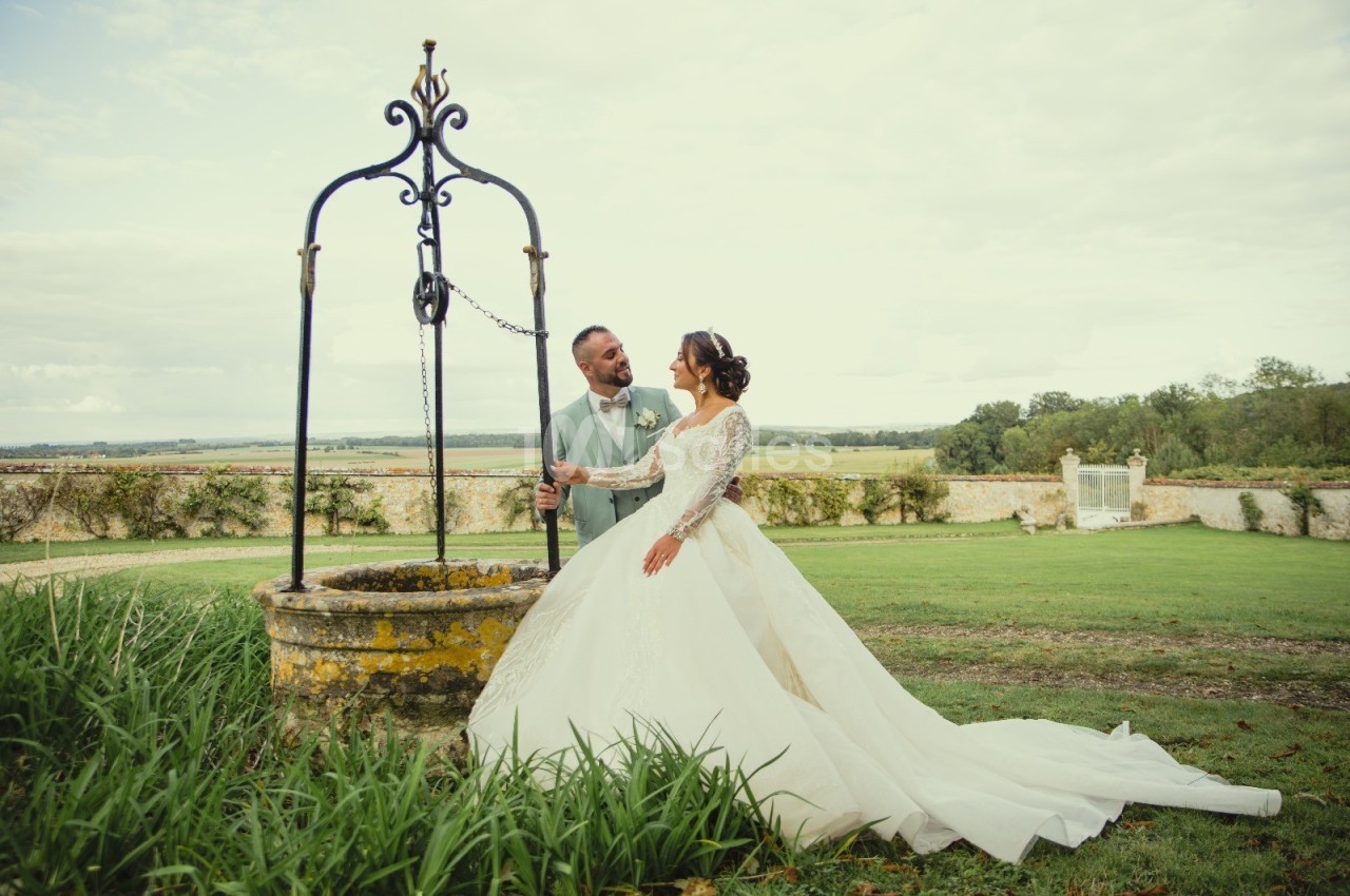 Un couple en tenue de mariage se tient près d'un puits ancien dans un jardin verdoyant.