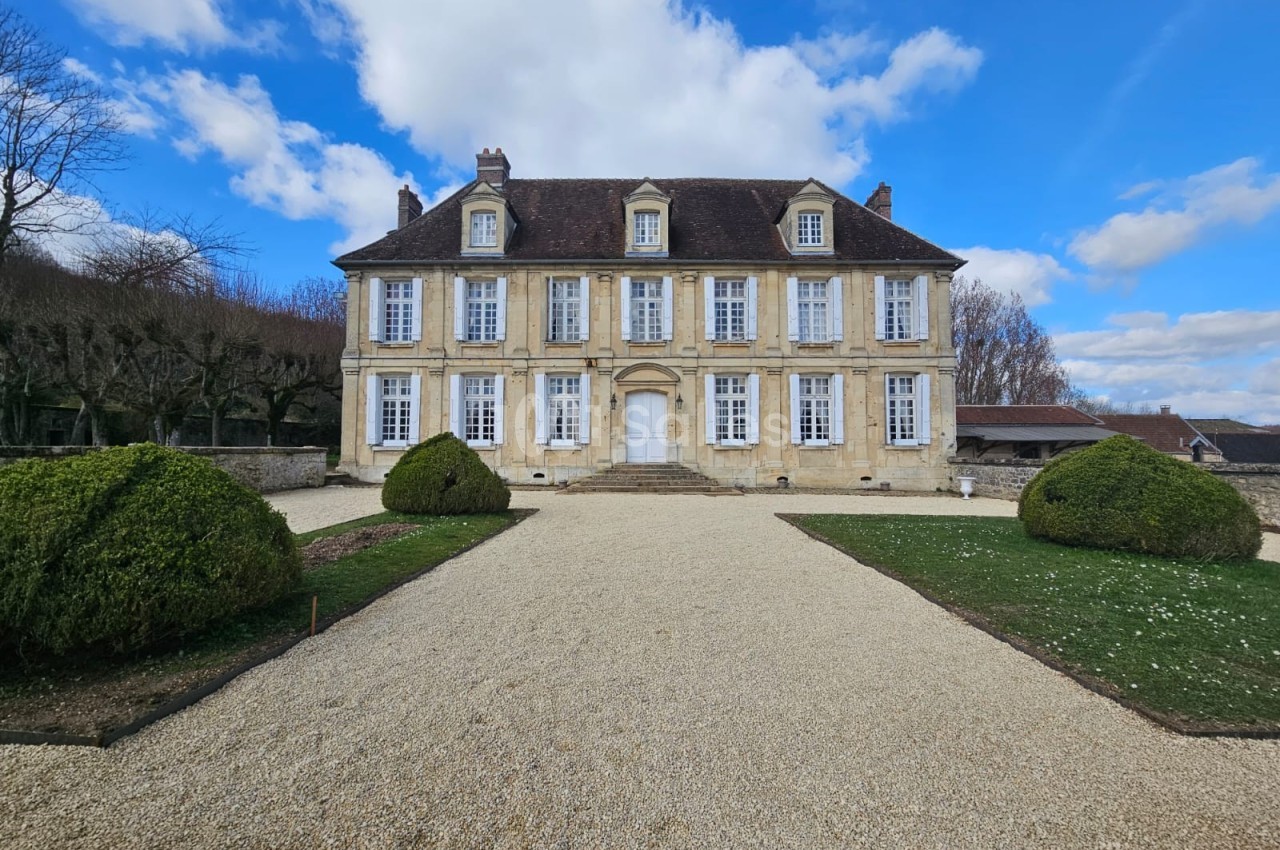 Façade d'un manoir en pierre avec toit en ardoise, entouré d'un jardin et d'une allée gravillonnée sous un ciel bleu.