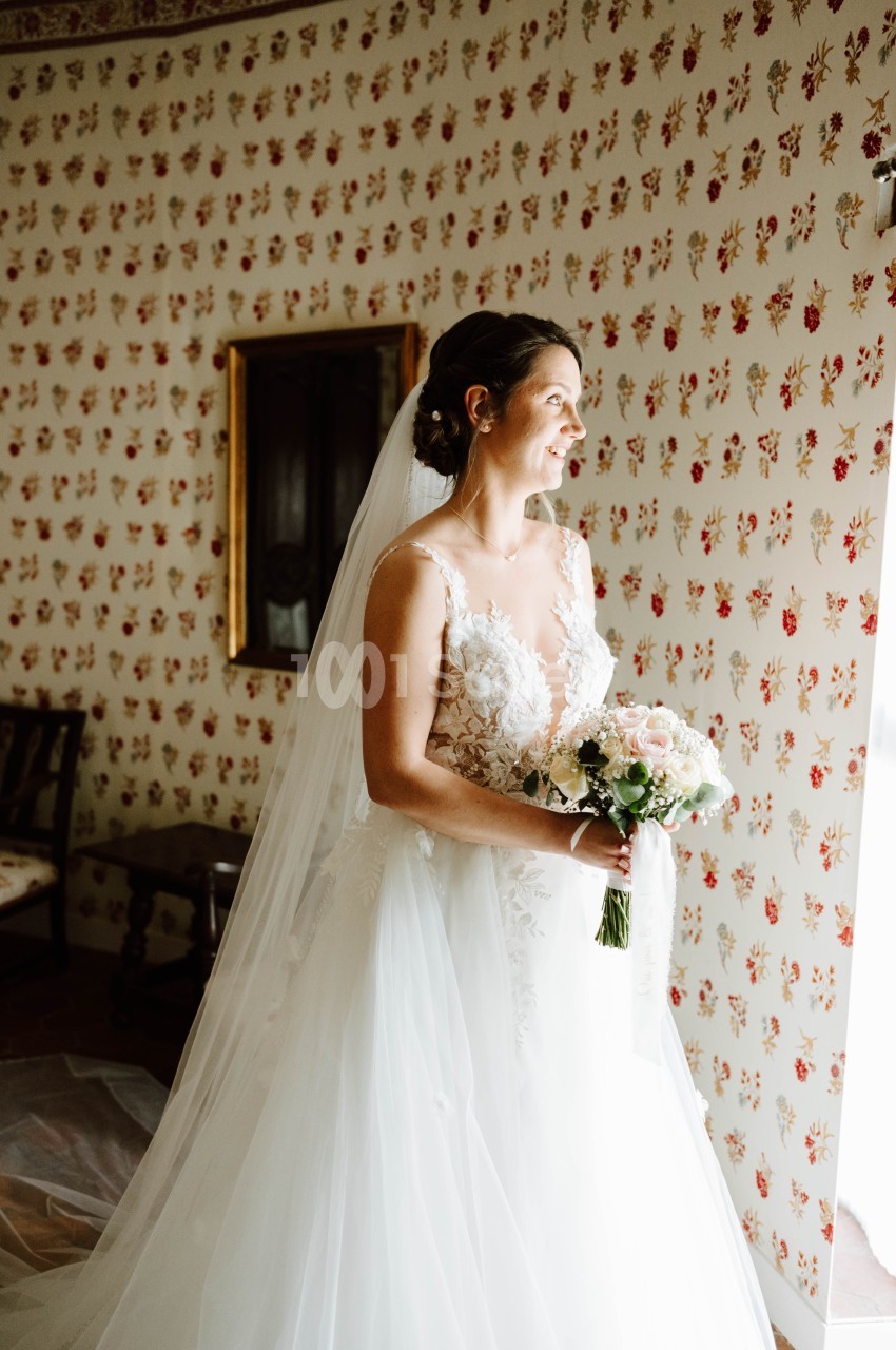 Une mariée en robe blanche tient un bouquet de fleurs, debout près d'une fenêtre dans une pièce au papier peint fleuri.