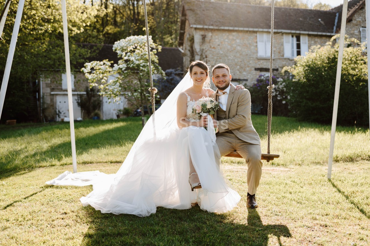 Un couple de mariés assis sur une balançoire dans un jardin, devant une maison en pierre sous un ciel ensoleillé.