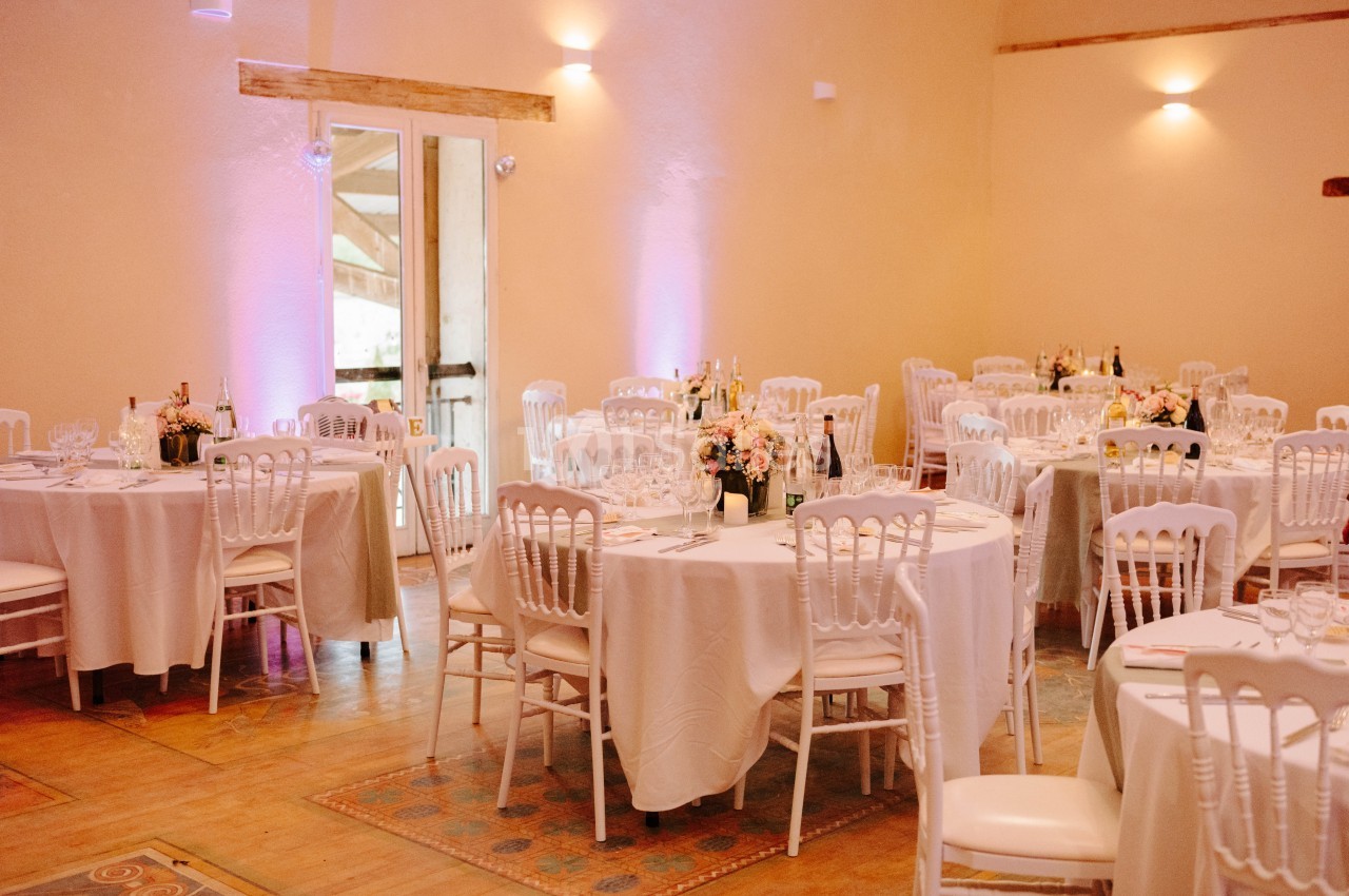 Salle de réception décorée avec des tables rondes, nappes blanches, chaises élégantes et arrangements floraux.