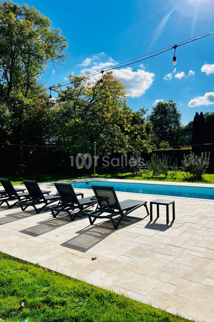 Chaises longues noires alignées près d'une piscine extérieure entourée de verdure sous un ciel ensoleillé.