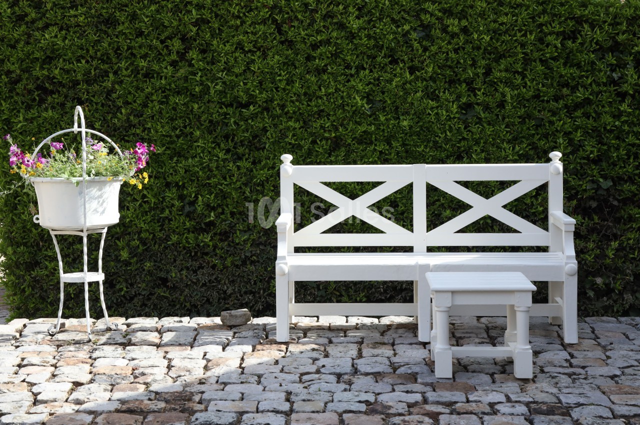 Banc et table blanche en bois sur une terrasse pavée, avec un support de fleurs blanches et un mur de haie verte.