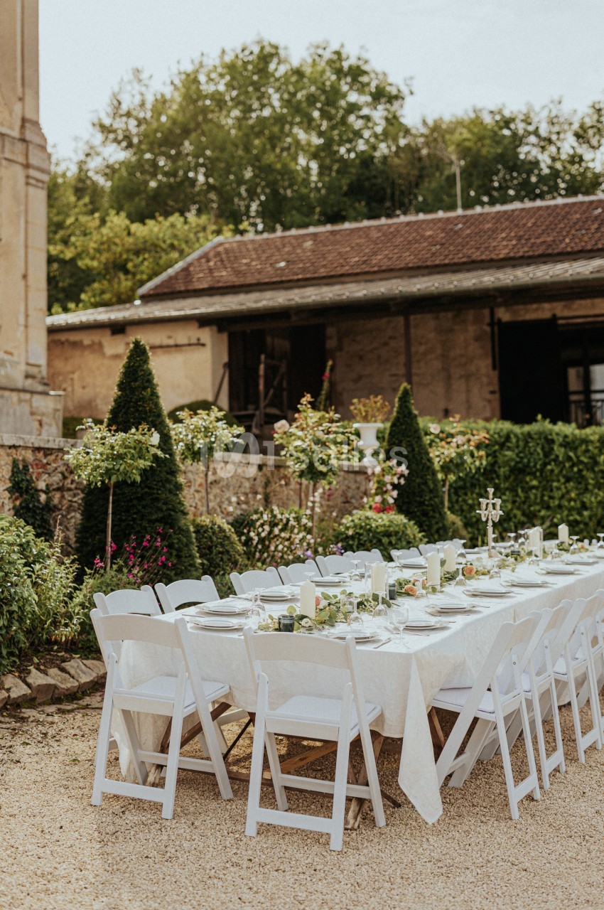 Table dressée en extérieur avec nappes blanches, chaises pliantes et décorations florales, devant un bâtiment ancien.