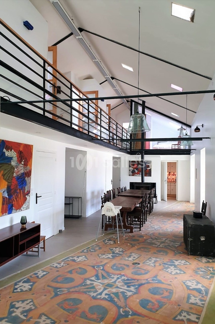 Intérieur lumineux avec mezzanine, grande table en bois, tapis coloré et œuvres d'art accrochées au mur.