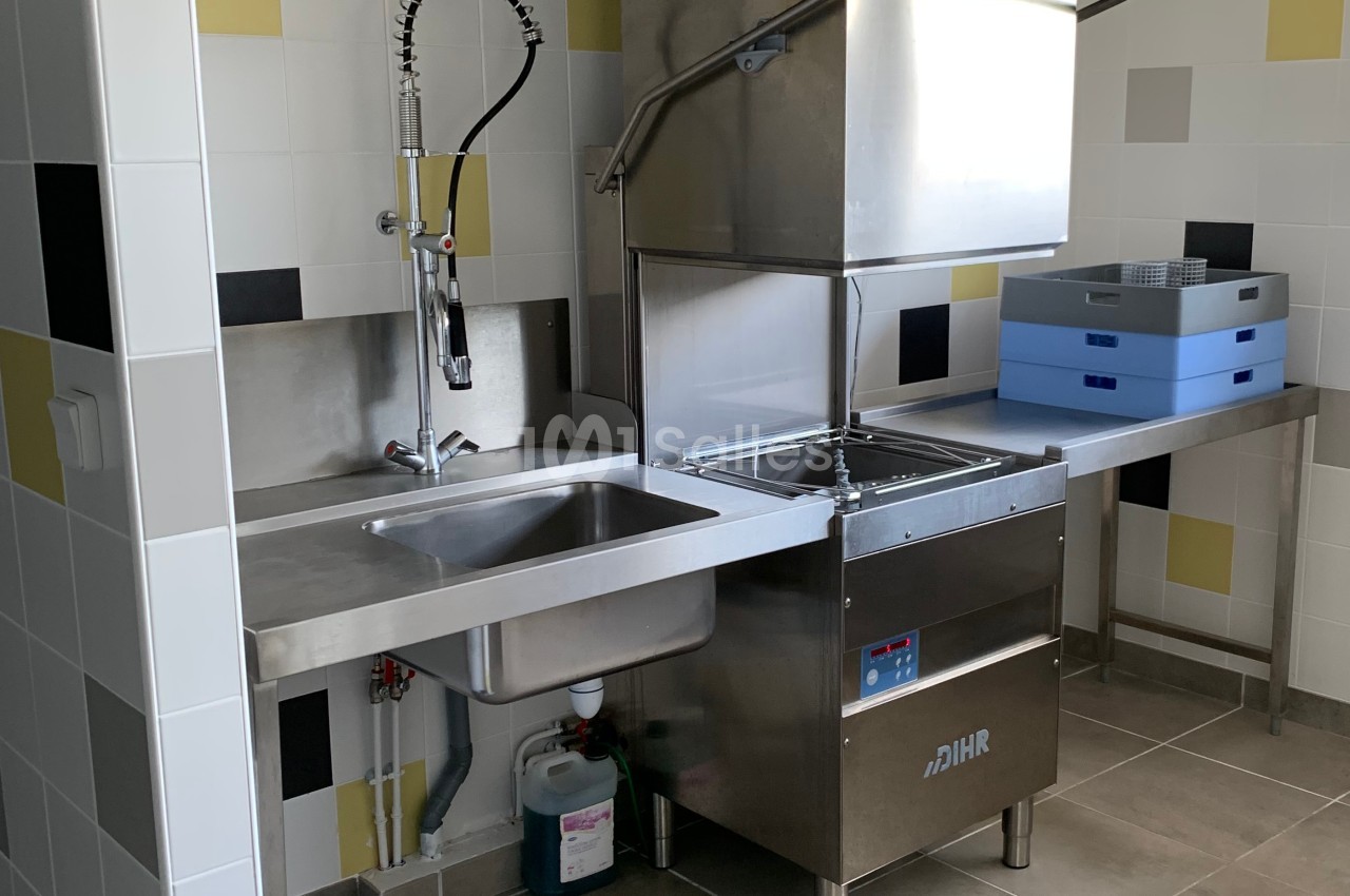 Évier en inox avec robinet professionnel, lave-vaisselle industriel et plans de travail dans une cuisine carrelée.